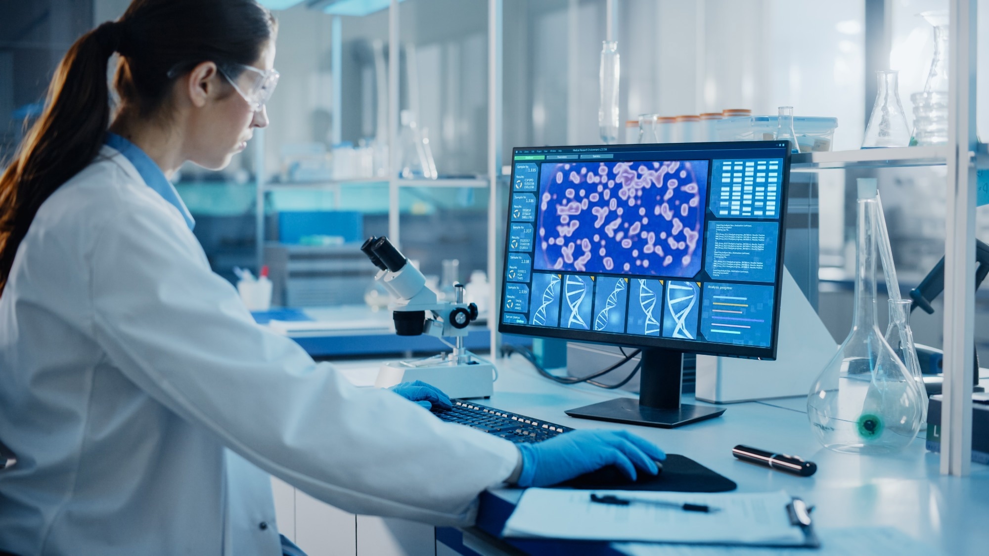 Female scientist in a lab coat and safety goggles analyzing DNA sequences and cell images on a computer monitor in a high-tech laboratory. Laboratory glassware and a microscope are visible on the desk.