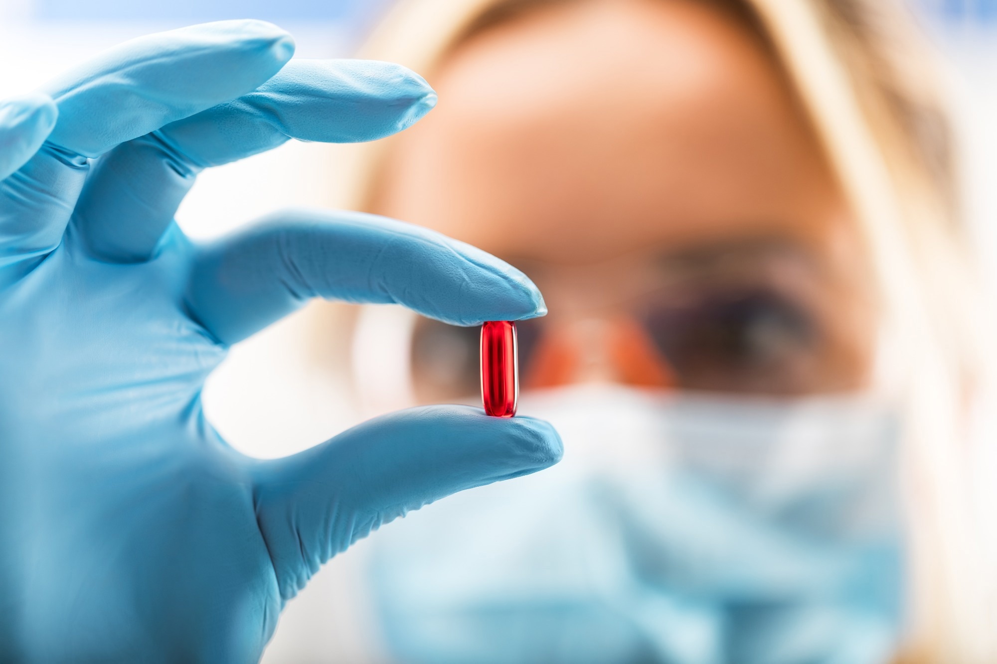 female scientist with protective eyeglasses and mask holding a red transparent pill with fingers in gloves in the pharmaceutical research laboratory