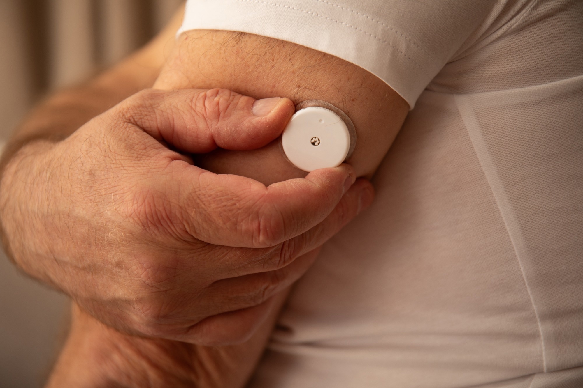 Man’s hands touching wearable glucose sensor on upper arm, neutral background with copy space on left
