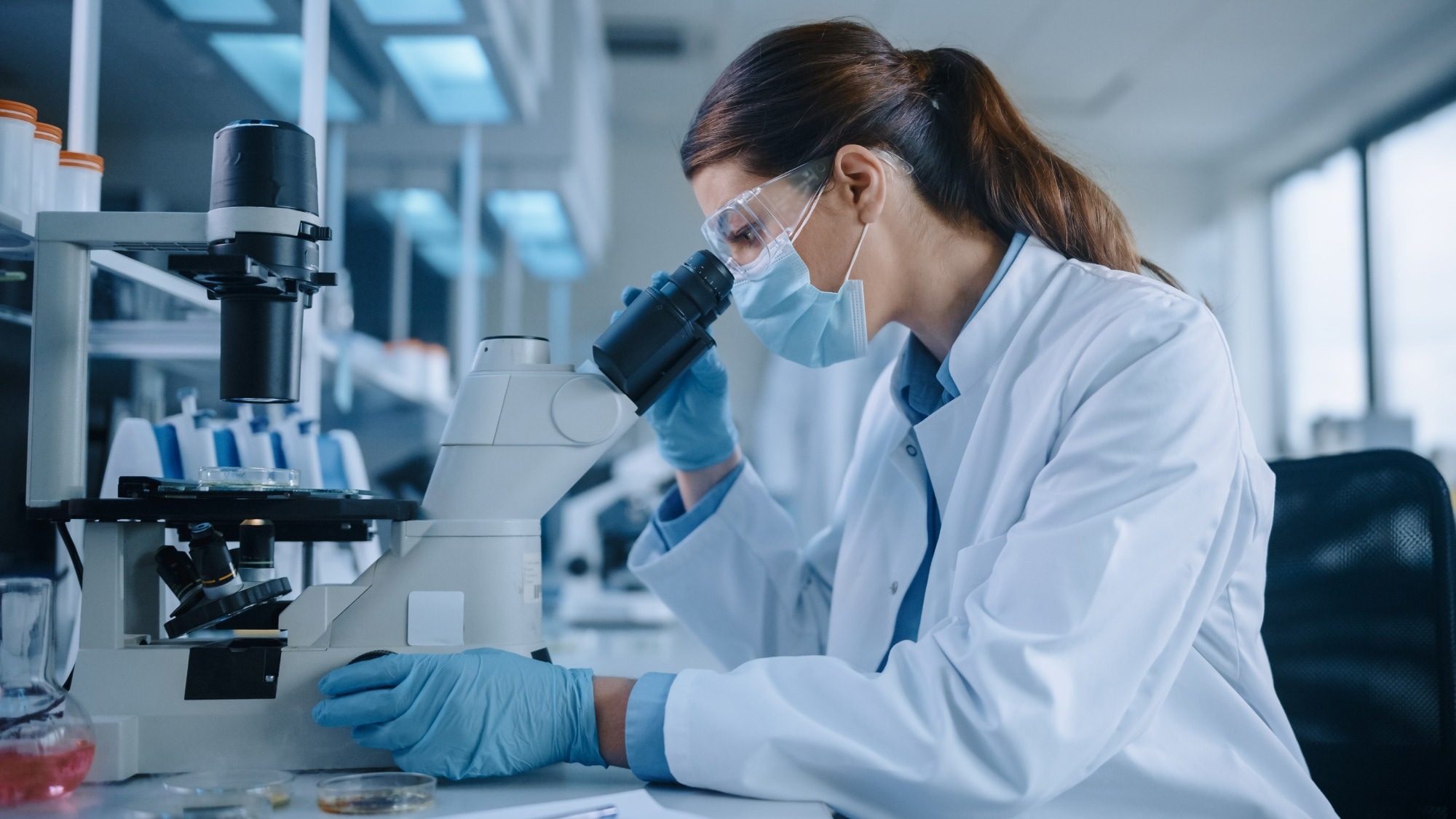 Female Scientist in Face Mask and Glasses Looking a Petri Dish with Genetically Modified Sample Chemicals Under a Microscope.